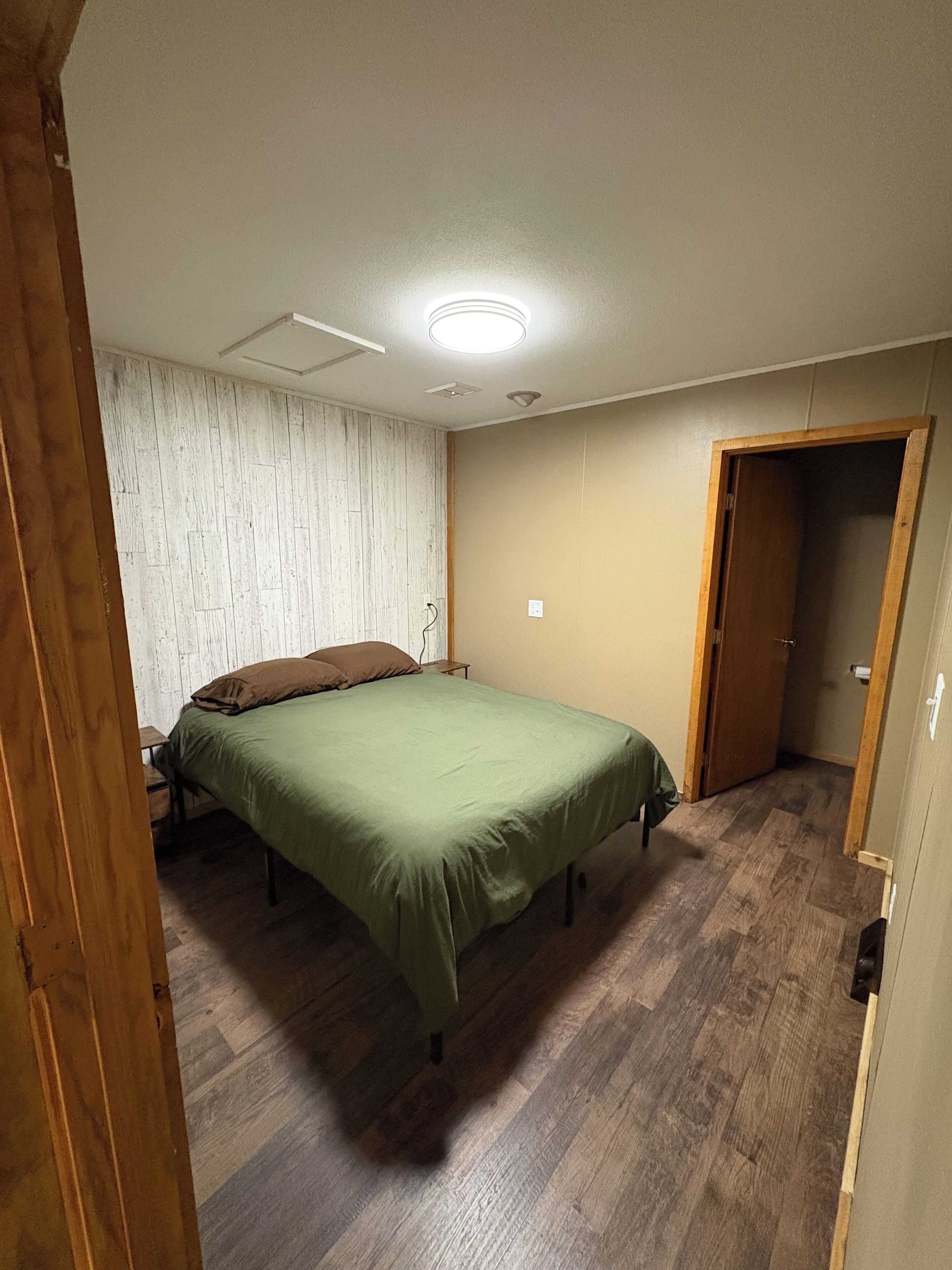 A bedroom with a wooden floor, a bed with a green bedspread, a wooden door, and a white wall with a textured pattern.