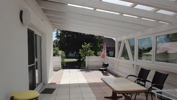 A covered patio area with a table and chairs, a grill, and a view of the garden. - Fewo Eichhölzle 1, 90 sqm, 2 Bedrooms, Max. 4 Guests (Münsingen)