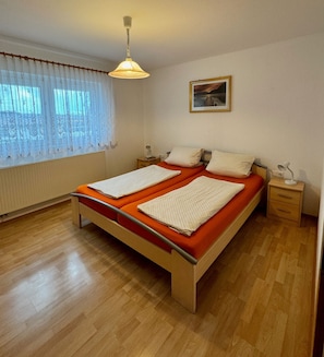 Una cama doble con cabecera de madera y ropa de cama naranja, dos mesitas de noche con lámparas, un ventanal con cortinas traslúcidas y un cuadro en la pared.