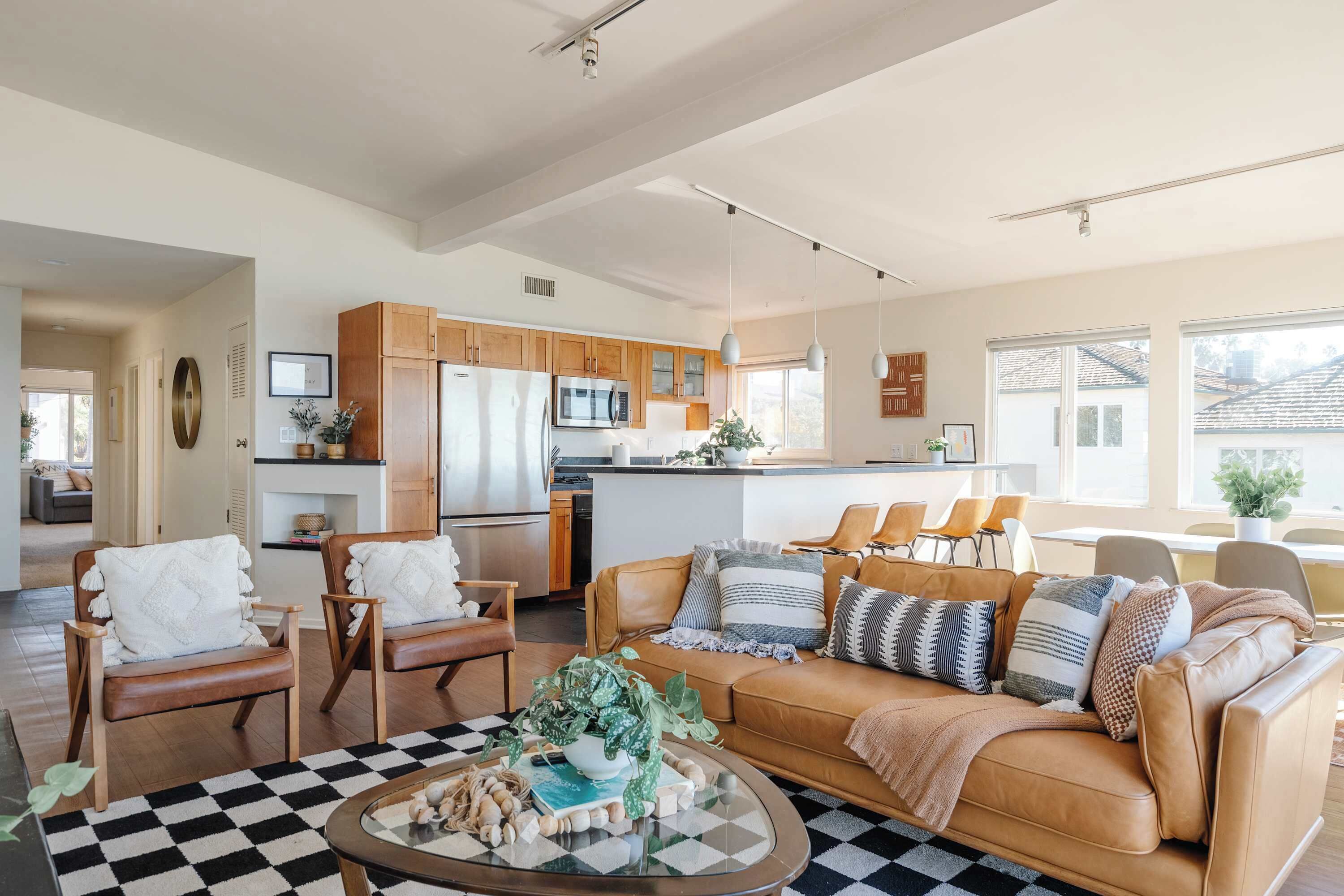 A spacious living room with a checkered rug, a tan leather sofa with multiple cushions, wooden chairs, and a coffee table with a plant.