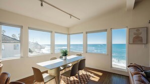 A modern dining area with a view of the ocean, a large window, and a brown leather couch. - Serene Escape at Windansea: Oceanview 2-Bedroom (San Diego)