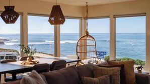 A room with large windows overlooking the ocean, wicker chairs, a dining table with a plant, and a hanging light fixture. - Beachfront Bliss: Oceanview Haven at Windansea (San Diego)