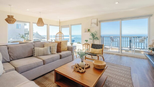 A spacious living room with a large gray sofa, a wooden coffee table, and a view of the ocean through large windows. - Beachfront Bliss: Oceanview Haven at Windansea (San Diego)