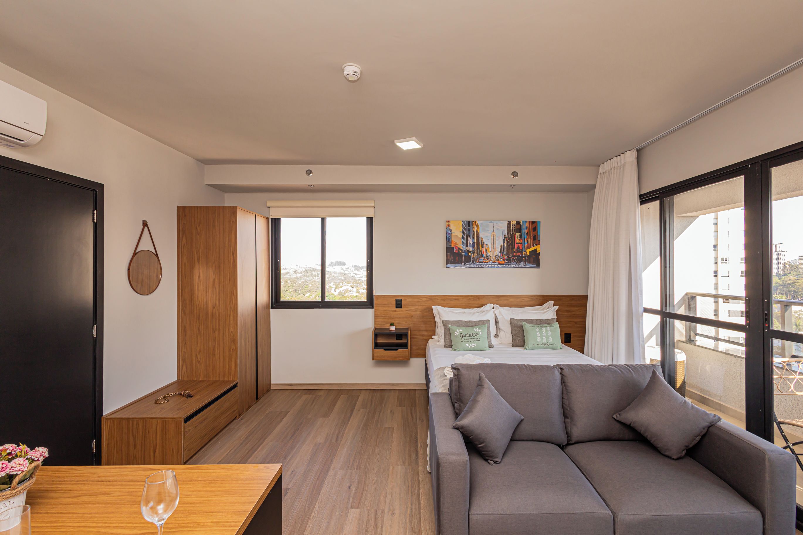 Une chambre d’hôtel moderne avec un lit, un canapé, une armoire en bois, un bureau avec un verre et un tableau au mur.