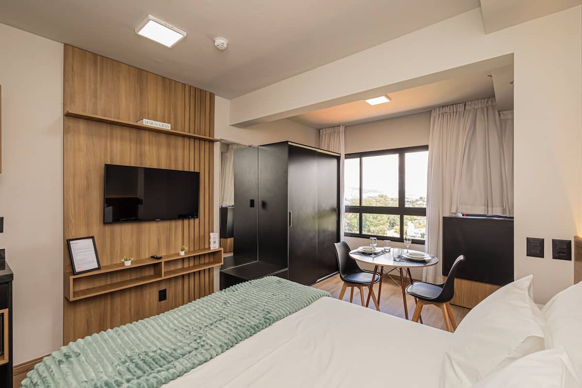 A modern hotel room with a bed, a desk with a chair, a television, and a large window with curtains.