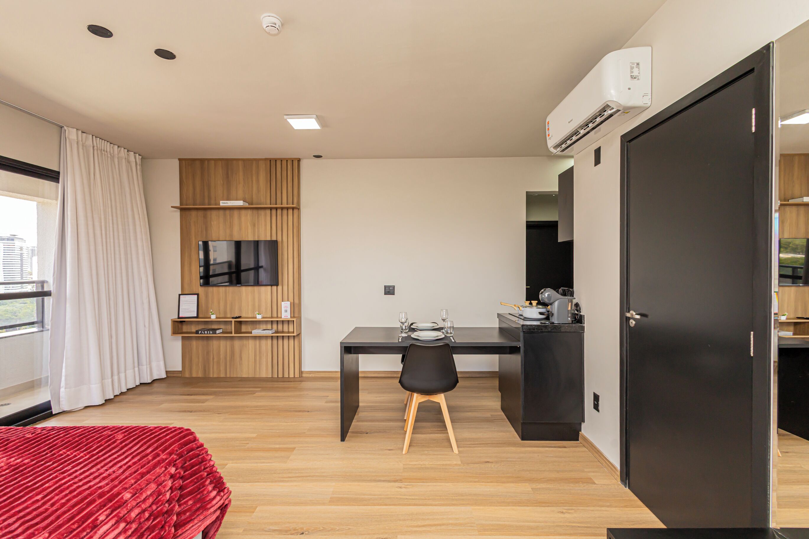 A modern hotel room with a wooden floor, a black desk with a chair, a TV mounted on the wall, and a large window with curtains.