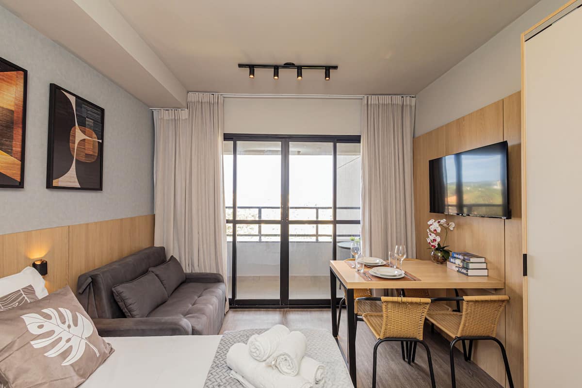 A hotel room with a bed, a sofa, a dining table with chairs, a TV, and a balcony with curtains.