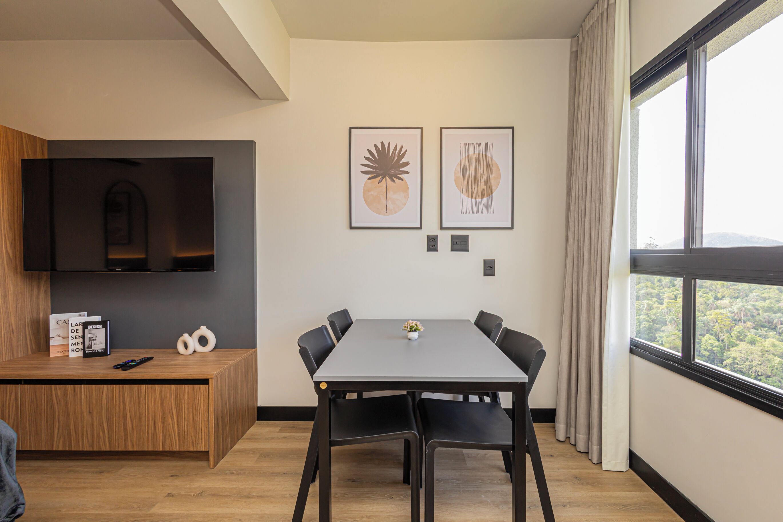 Une chambre moderne dotée d’une télévision à écran plat, d’un bureau en bois, d’une table à manger avec des chaises et d’œuvres d’art encadrées accrochées au mur.