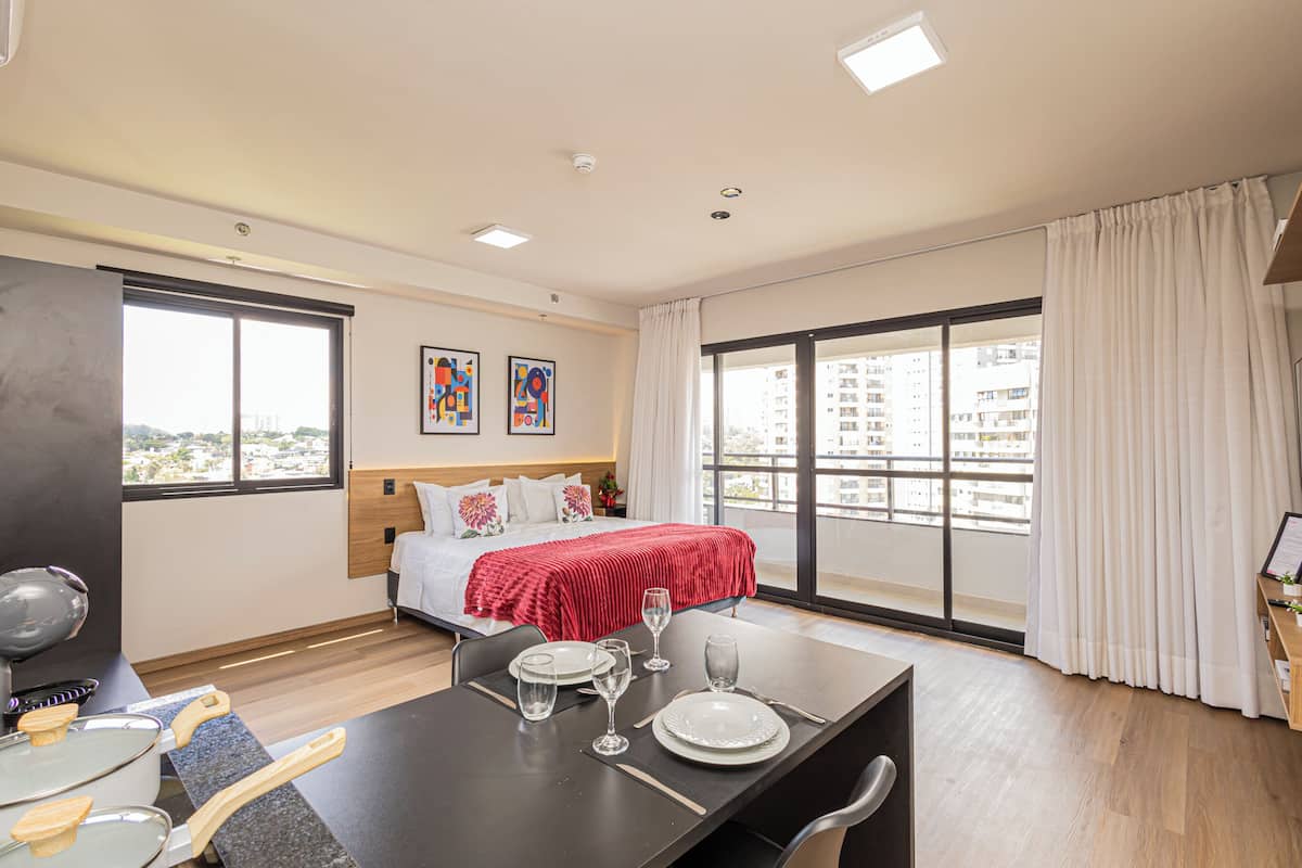 A modern hotel room with a bed, a dining area, and a large window with a city view.