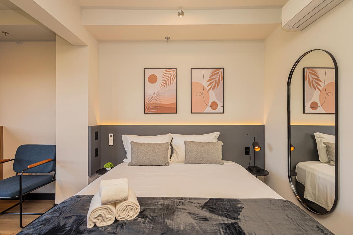 A hotel room with a bed, a chair, a wall-mounted mirror, and two framed artworks.