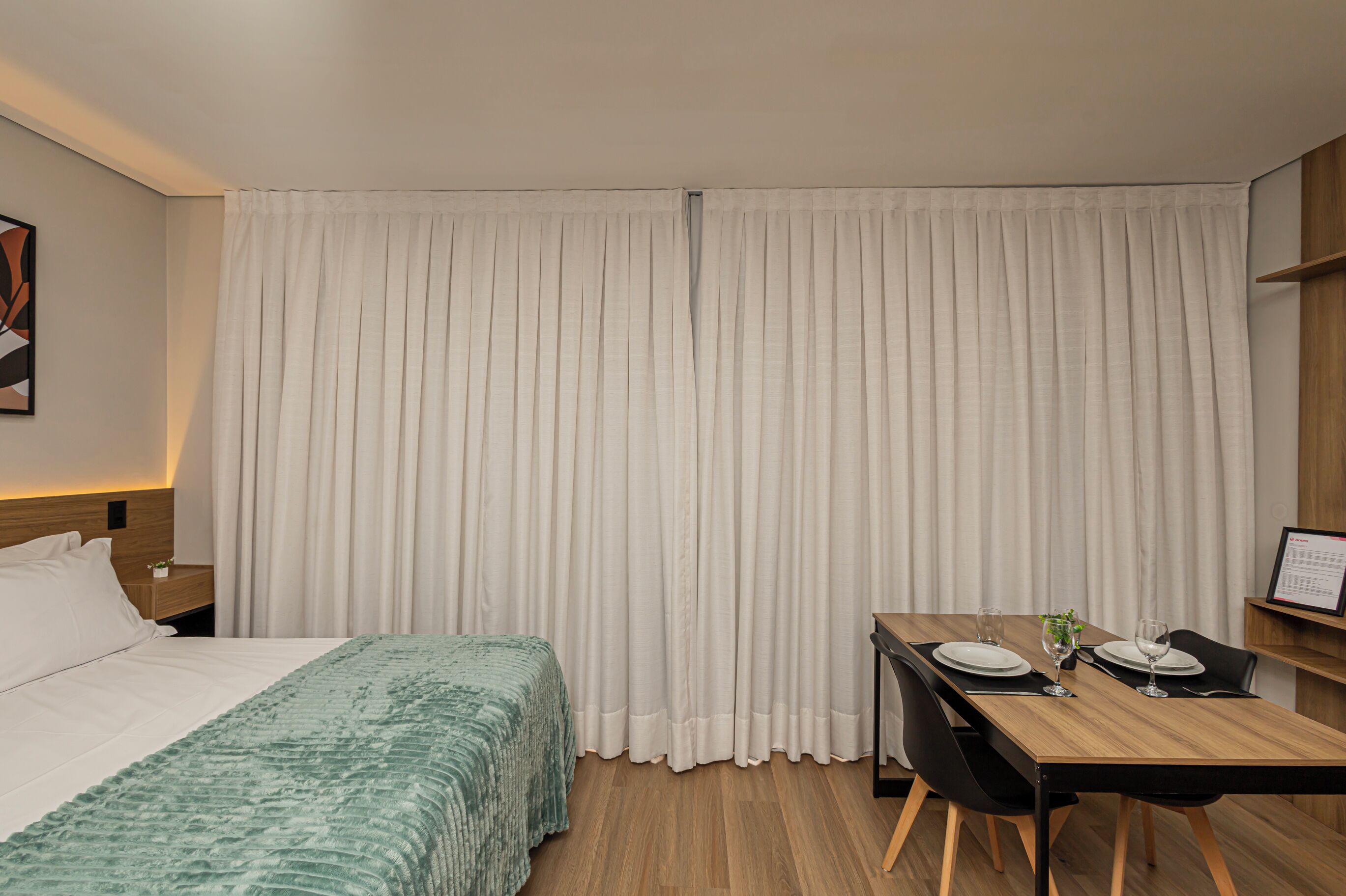 A hotel room with a bed, a wooden desk with two chairs, a small table with a glass and a plant, and a white curtain covering the window.