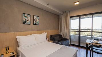 Una habitación de hotel con cama, un escritorio con silla, una ventana con cortinas y una obra de arte en la pared.
