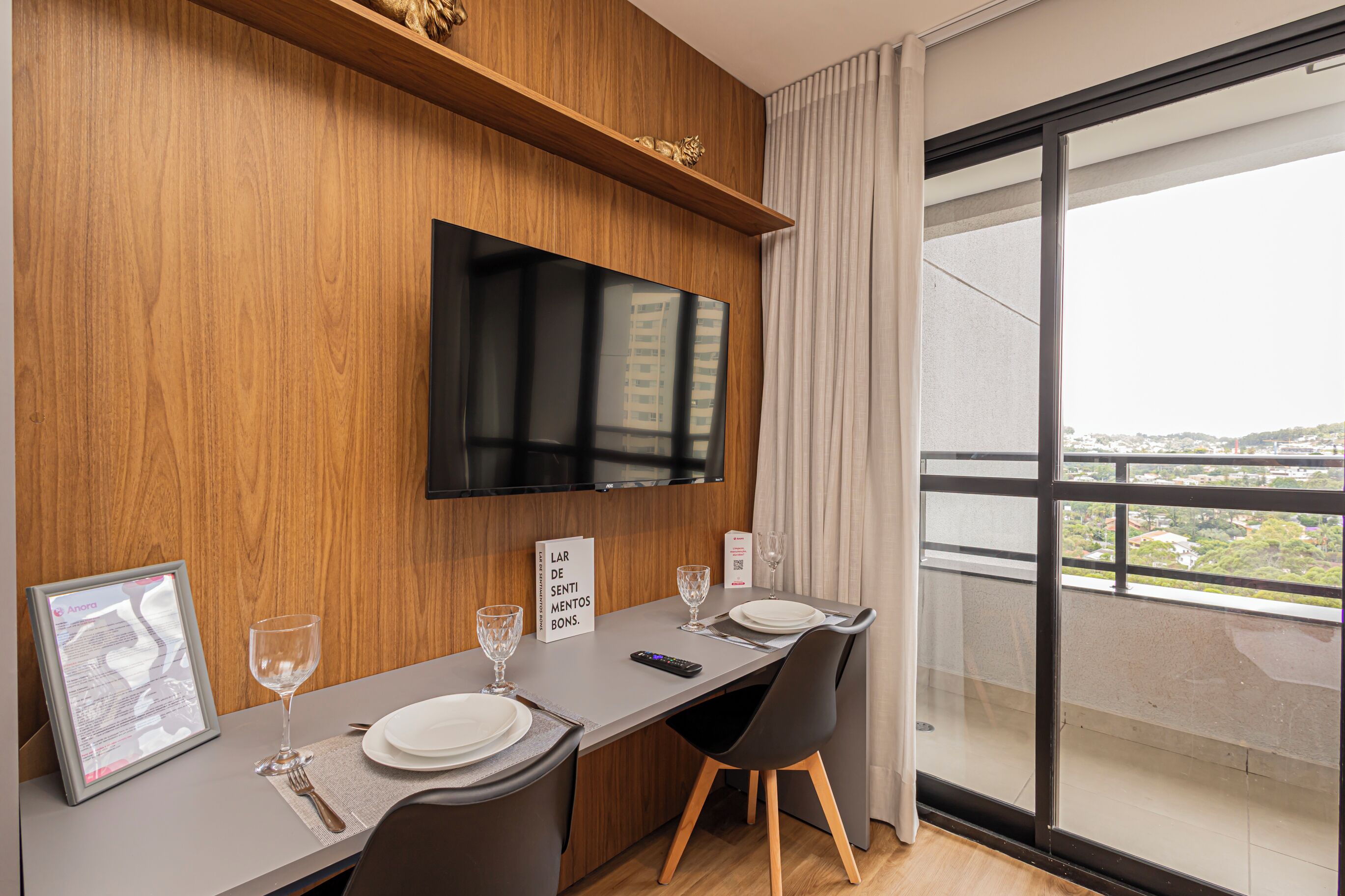 Ein modernes Hotelzimmer mit einer Holzwand, einem Flachbildfernseher, der über einem Esstisch angebracht ist. Der Tisch ist mit weißen Tellern, Gläsern und Besteck gedeckt. Eine eingerahmte Speisekarte liegt auf dem Tisch, und eine Fernbedienung ist sichtbar. Das Zimmer hat ein großes Fenster mit Blick auf die Stadtlandschaft.