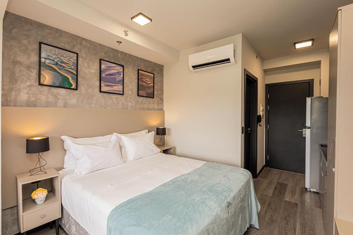 A hotel room with a bed, nightstands, a lamp, an air conditioner, and a door.
