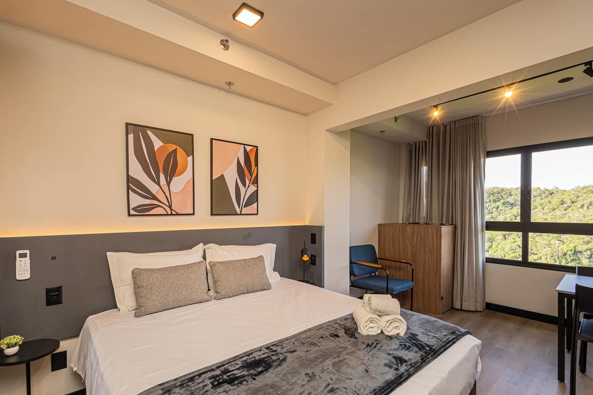 A modern hotel room with a large bed, a desk, a chair, and a view of the outdoors.