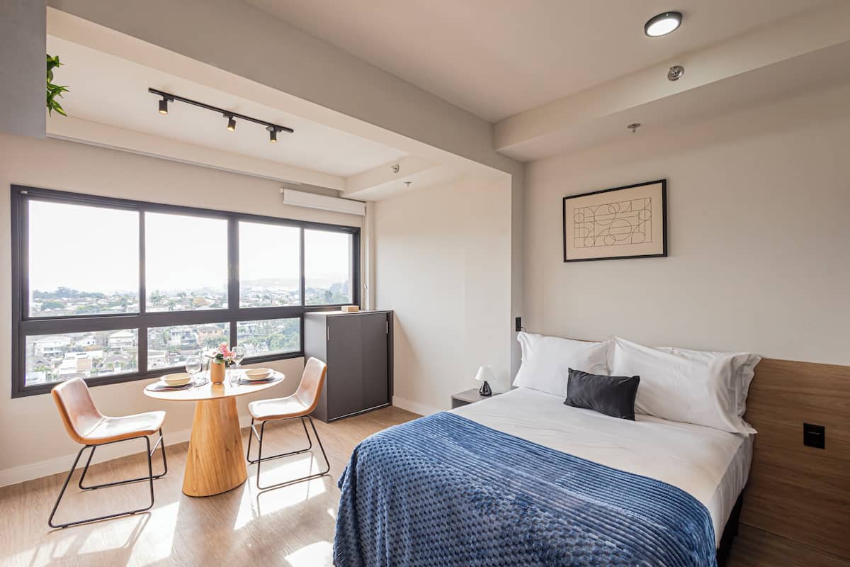 A modern hotel room with a large window, a round dining table, two chairs, a bed with a blue blanket, and a small bedside table with a lamp.