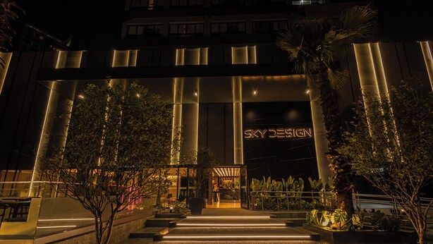 Ein moderner Hotelzugang mit einem gut sichtbaren Schild mit der Aufschrift "Sky Design", einem gut beleuchteten Weg zum Eingang und umliegenden begrünten Außenanlagen mit verschiedenen Pflanzen.