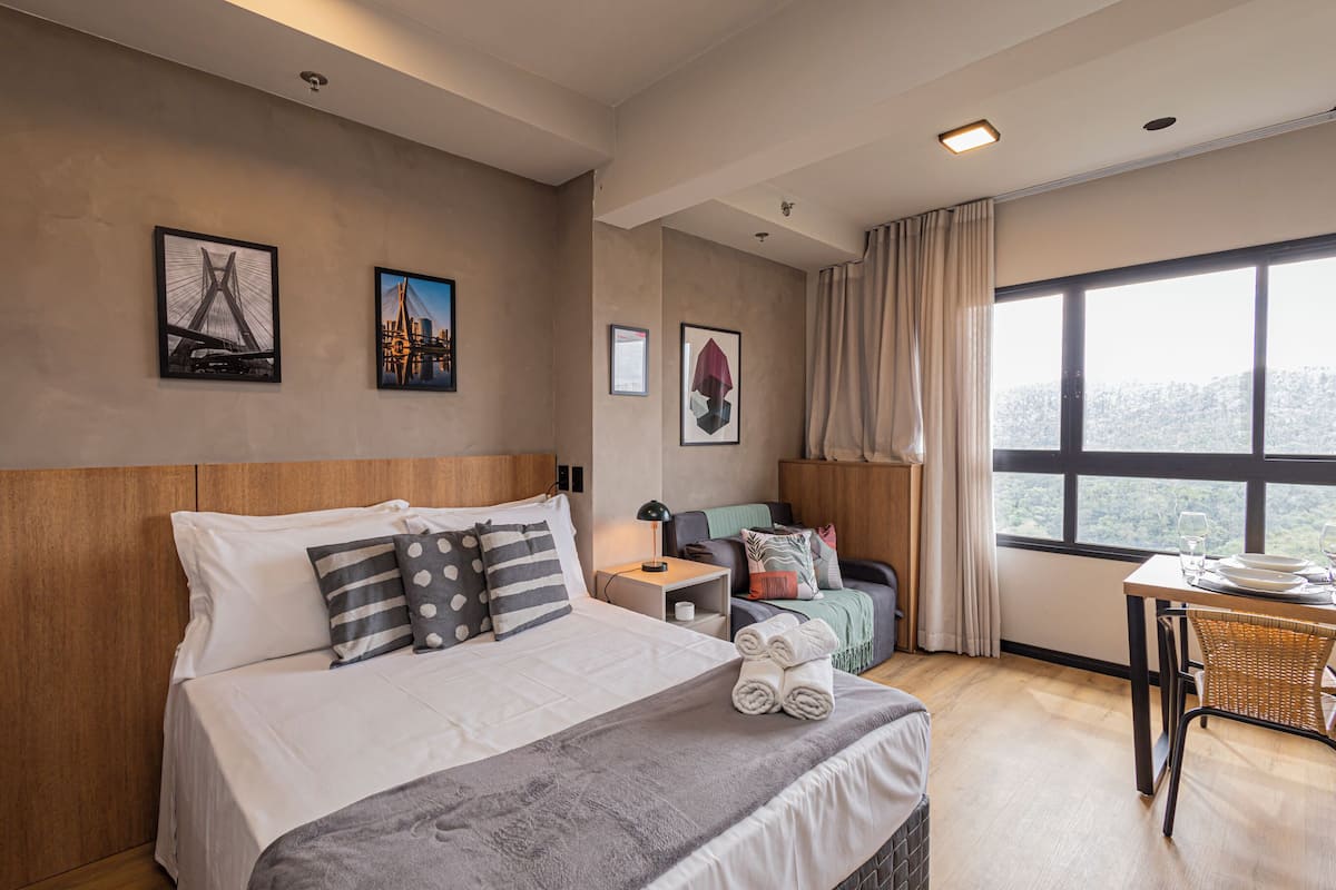 A hotel room with a bed, a nightstand, a table with a chair, and a large window with a view.