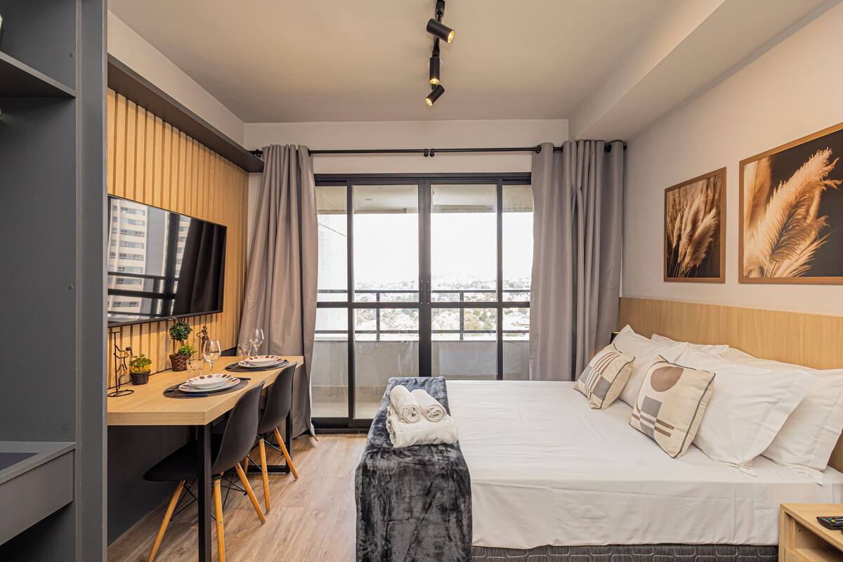 A modern hotel room with a large bed, a desk with a TV, and a balcony with curtains.