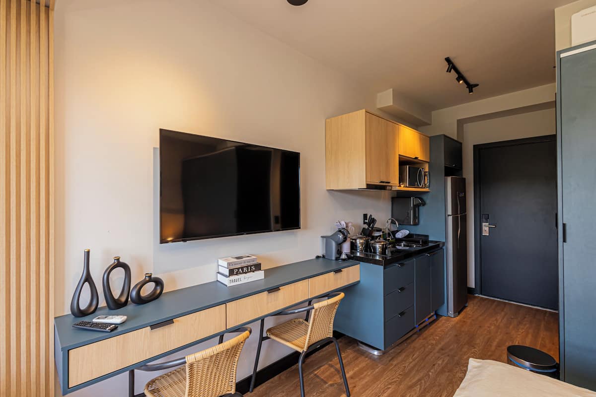 A compact hotel room with a flat-screen TV, a kitchenette, a dining area with chairs, and a bed.