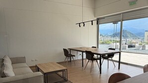 Moderni olohuone, jossa on sohva, sohvapöytä ja ruokailutila tuoleineen. Suuri ikkuna avautuu kaupunkinäkymään.