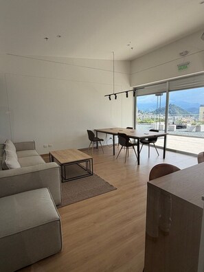 Un salon moderne avec un canapé, une table basse et un coin repas avec des chaises. Une grande fenêtre offre une vue sur la ville.