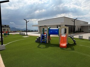 An outdoor play area with a slide and climbing structures, surrounded by artificial grass and a building in the background. - Comfortable & Stylish – Premium Area (Manizales)