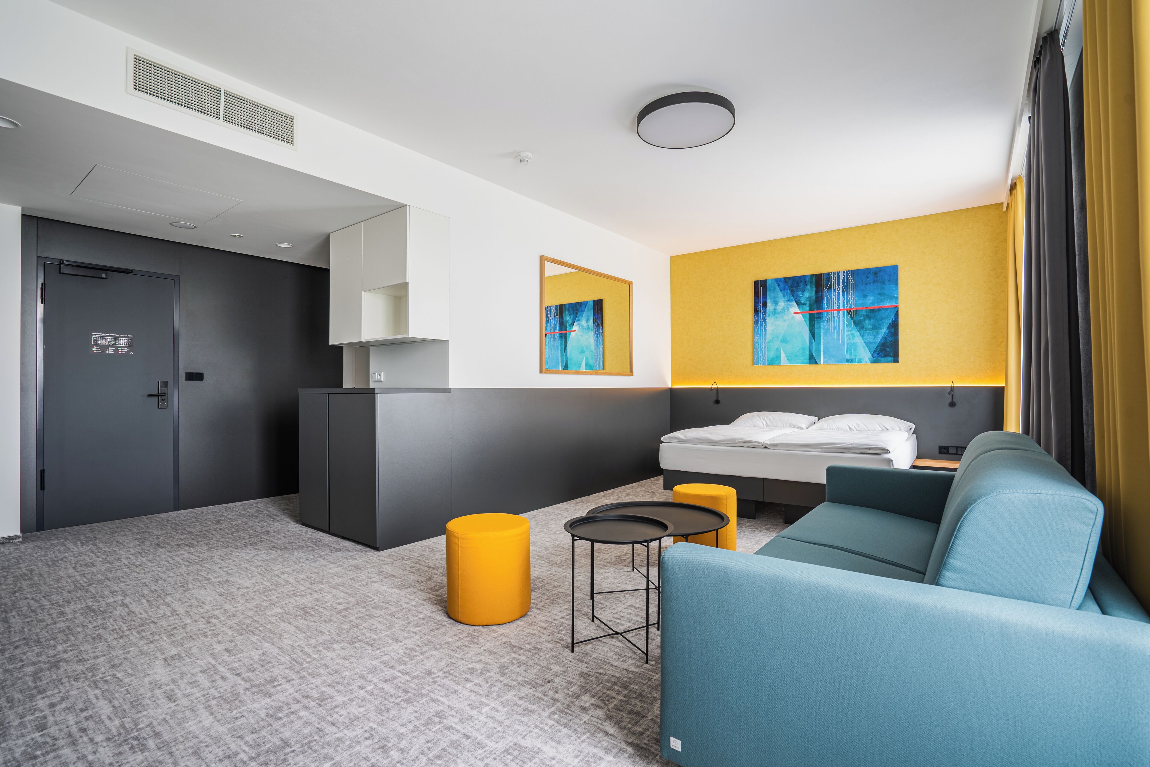 A modern hotel room with a bed, a sofa, a small table, and a yellow ottoman.