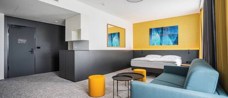A modern hotel room with a bed, a sofa, a small table, and a yellow ottoman.