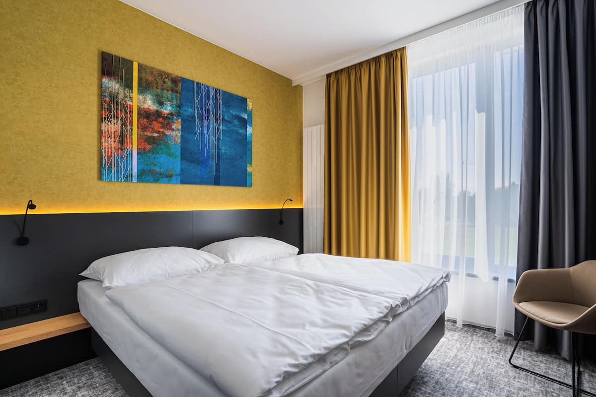 Een moderne hotelkamer met een groot bed, nachtkastje, stoel, schilderij aan de muur en raam met gordijnen.