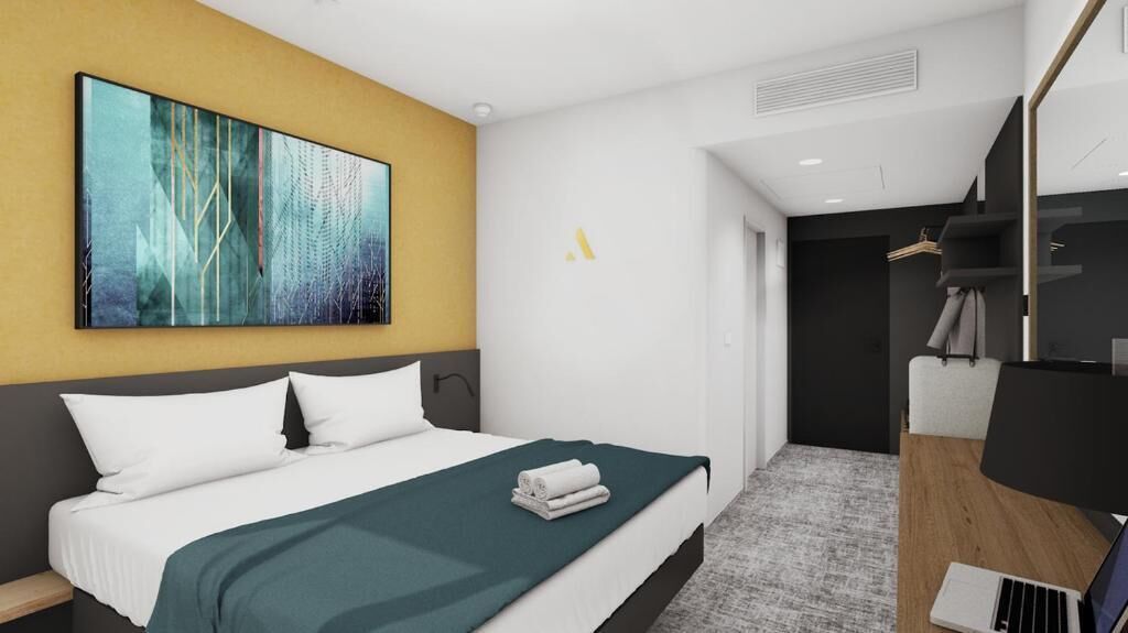 A modern hotel room with a large bed, a desk with a laptop, a painting on the wall, and a black wardrobe.