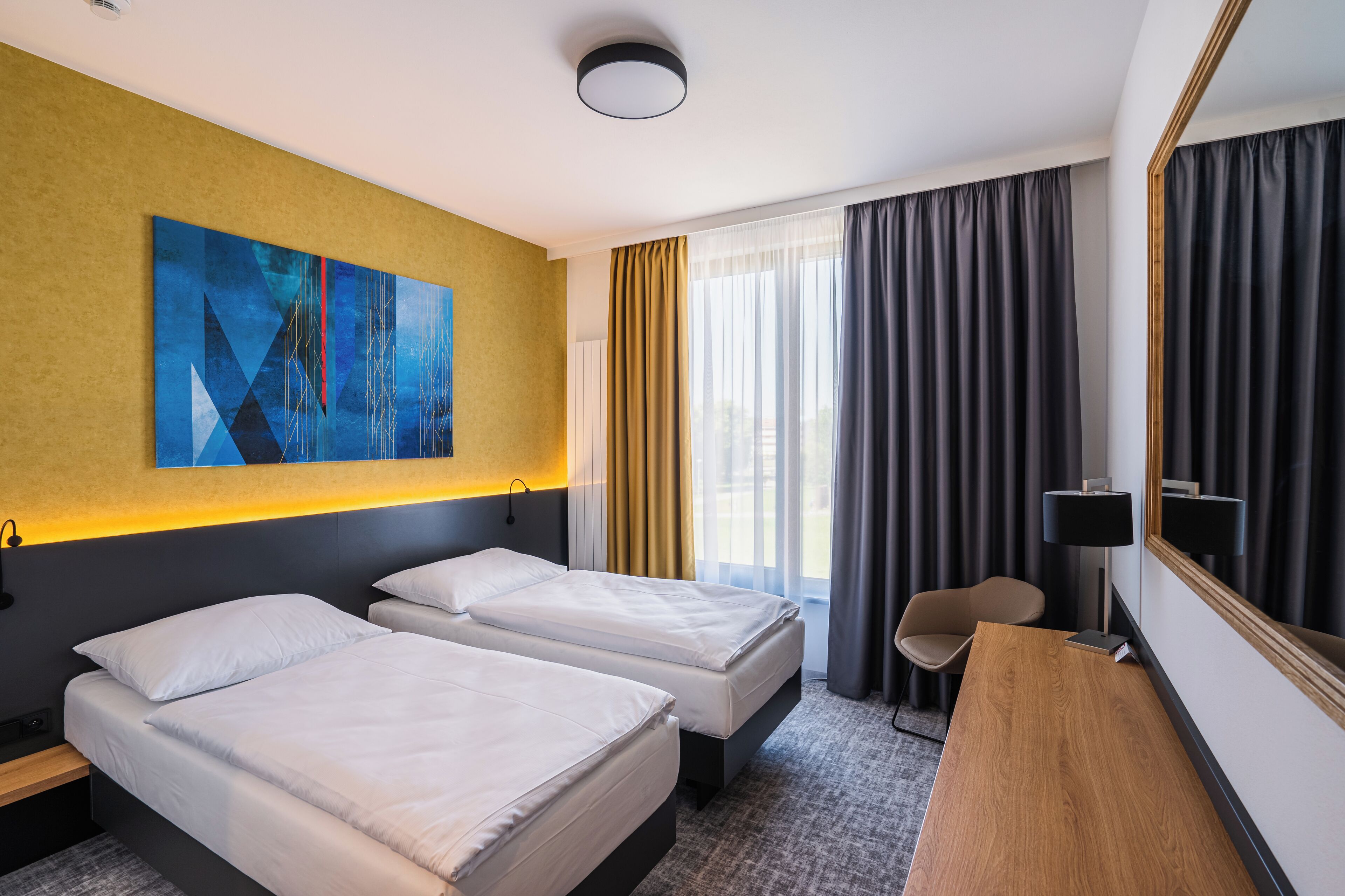 A hotel room with two beds, a desk, a chair, a large mirror, and a painting on the wall.