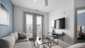 A modern living room with a glass coffee table, a sofa, a desk, and a flat-screen TV mounted on the wall. - Landing | Modern 1BD, Gym, Pool (Nashville)