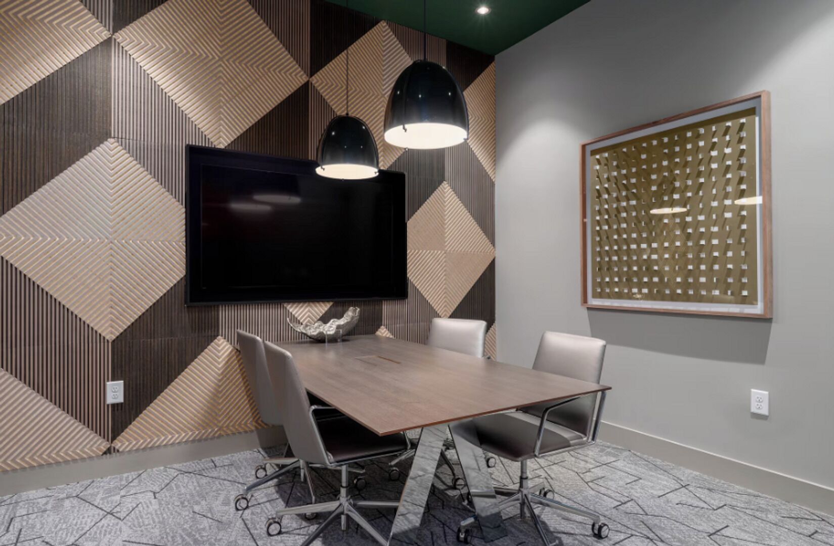 A conference room with a large flat-screen TV, two pendant lights, a wooden table, four chairs, and a decorative wall panel.