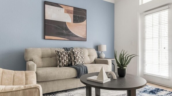 Ein Wohnzimmer mit einem beigefarbenen Sofa, gestreiften Kissen, einem runden Couchtisch mit einem Buch, einer Topfpflanze und einem großen abstrakten Gemälde an der Wand.