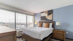 Camera da letto moderna con un letto grande, testiera in legno e vista sulla città attraverso ampie finestre a tutta altezza.