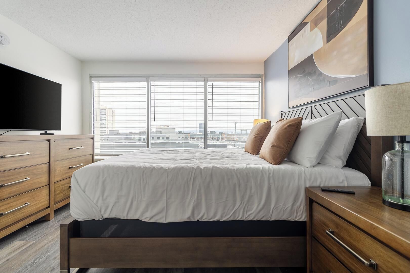 Une chambre d’hôtel moderne dotée d’un grand lit, de tables de chevet en bois, d’une télévision et offrant une vue sur la ville grâce à de grandes fenêtres du sol au plafond.