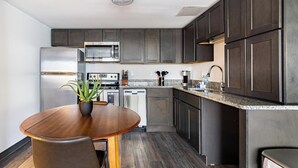 A modern kitchen with dark cabinets, stainless steel appliances, a granite countertop, and a wooden dining table with a potted plant. - Landing | Stylish 1BD, Yoga Studio, Game Room (Hartford)