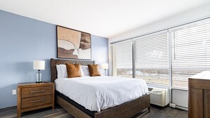 A modern hotel room with a large bed, wooden headboard, and a view of the city through floor-to-ceiling windows. - Landing | Stylish 1BD, Yoga Studio, Game Room (Hartford)