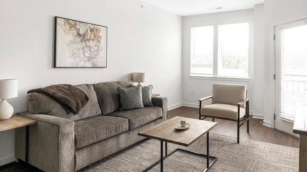 A modern living room with a grey sofa, a wooden coffee table, a chair, and a painting on the wall. - Landing | Incredible 1BD, Gym, Clubhouse (Raleigh)