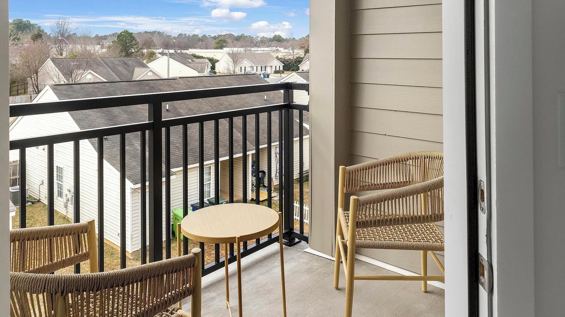 Varanda com mesa e duas cadeiras de vime, com vista para uma área residencial com casas e céu claro.