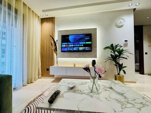 Ein modernes Wohnzimmer mit flachbildfernseher an der Wand, einem Marmortisch, einer Fernbedienung, einer Vase mit Blumen und einer Topfpflanze.
