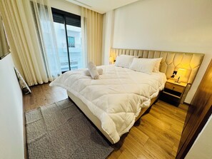 Ein Hotelzimmer mit einem großen Bett, einem Nachttisch mit Lampe, einem Fenster mit Vorhängen und Blick auf ein Gebäude.