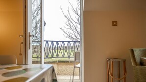 Ein Zimmer mit einer Tür, die auf einen Balkon führt, einem Tisch mit gemustertem Tischlauf, einem Stuhl und einem kleinen hölzernen Hocker.