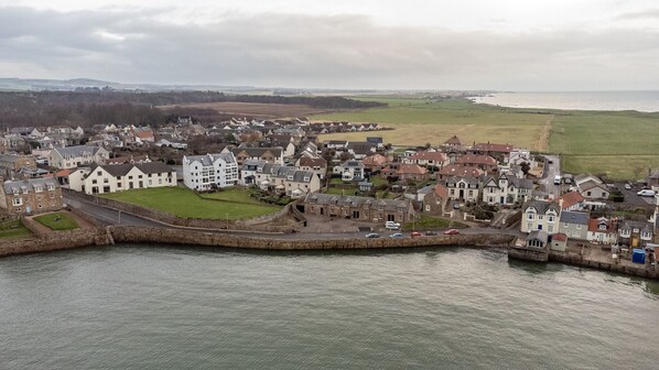 An aerial view of a coastal town with a mix of residential buildings, a prominent white building with multiple floors, and a stone wall along the waterfront. - Burnside, The Toft | Elie (Elie)