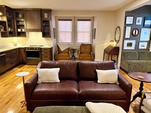 Un salon avec un canapé en cuir marron, une petite table ronde, un fauteuil vert et un coin cuisine doté de meubles en bois et d’une cuisinière.