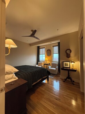 Une chambre à coucher avec un lit, deux chaises, une table avec des lampes, un ventilateur de plafond et une horloge au mur.