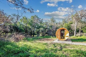 A wooden cylindrical structure with an open door, surrounded by a natural landscape with trees and grass.