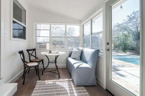 A sunroom with a pool view, a seating area with a sofa and chairs, and a small table with a lamp.
