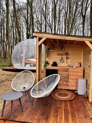 A wooden cabin with a transparent dome, a kitchen area, and two white wicker chairs on a wooden deck.